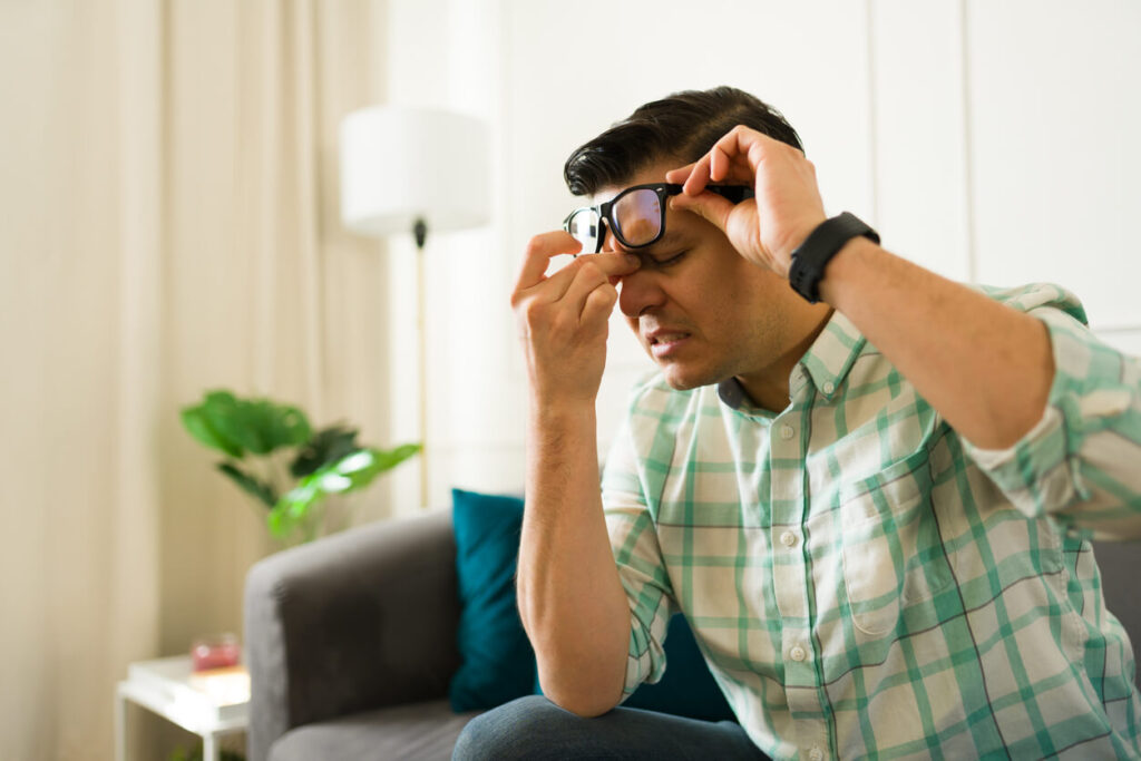 A man removing glasses, pressing nose bridge while experiencing intense headache on living room couch