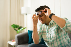 A man removing glasses, pressing nose bridge while experiencing intense headache on living room couch